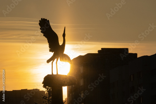 Urban landscape. Sculpture of a stork in a nest between high-rise buildings on the background of the setting sun. Contour image. Selective focus.