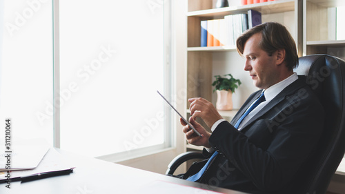 Professional businessman concentrate and culculating looking down to his tablet at office desk next to the window with copy space to the left, concept contemplative employer, successful entrepreneur.