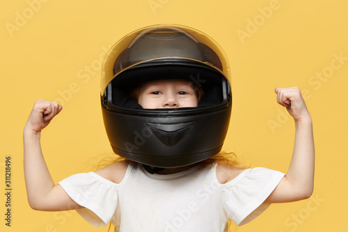 Fotografia Isolated shot of little girl racer posing against yellow studio wall background wearing black safety motorcycle helmet demonstrating her bicep muscles