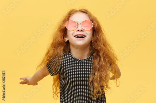 Children, style and fshion concept. Carefree fashionable little girl with curly red hair having happy joyful facial expression, laughing, wearing stylish pink sunglasses, keeping arms behind her back photo