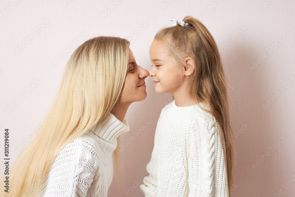 Side view of happy young female with long blonde hair going to kiss her charming little daughter posing with tips of noses pressed against each other. Love, family, generations and relationships