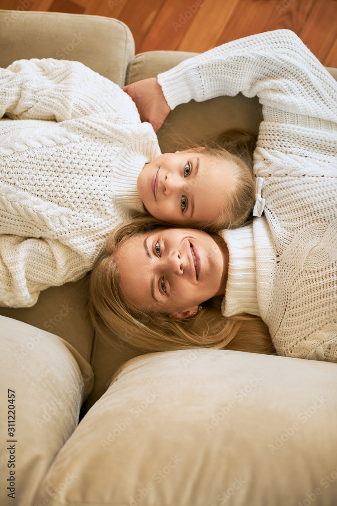 Top view of happy family relaxing at home. Vertical shot of beautiful young mother and her cute daughter lying head to head next to each other on sofa, smiling happily at camera, wearing sweaters Top view of happy family relaxing at home. Vertical shot of beautiful young mother and her cute daughter lying head to head next to each other on sofa, smiling happily at camera, wearing sweaters