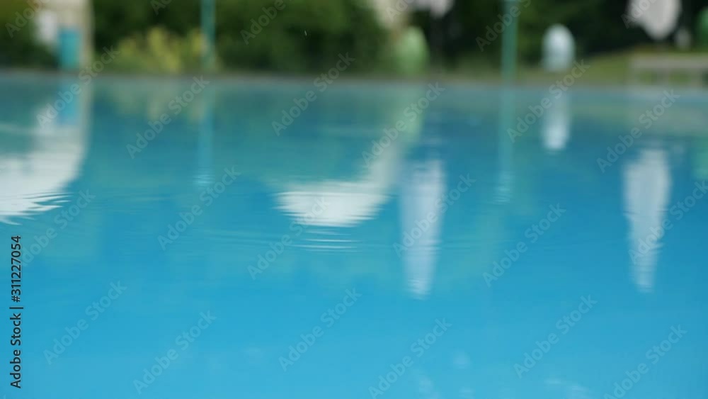 Rain drops fall on a surface of a swimming pool in slow motion. Rain splashing into a blue pool. Close up of swimming pool during storm. Rain drops fall into water. Rain drops fall on a surface of a swimming pool in slow motion. Rain splashing into a blue pool. Close up of swimming pool during storm. Rain drops fall into water.