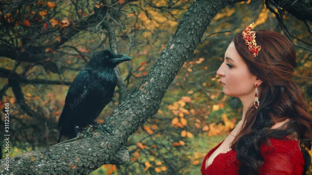 Close-up portrait of queen stroking touching big raven with tenderness. wild bird sits on branch of autumn tree with yellow leaves. Hair flying in wind. Gold crown jewelry. Backdrop fabulous nature