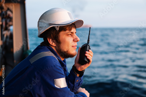 Marine Deck Officer or Chief mate on deck of offshore vessel or ship , wearing PPE personal protective equipment - helmet, coverall. He holds VHF walkie-talkie radio in hands.