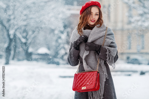 Happy smiling woman enjoying winter holidays, posing in snow covered street of European city. Model wearing beret, grey scarf, coat, gloves, with red quilted bag. Copy, empty space for text 