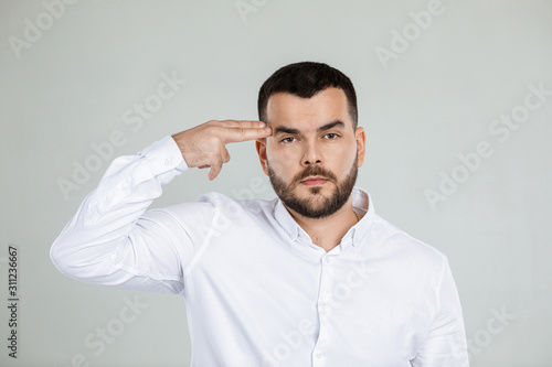 bearded man in white shirt shooting in temple with hand on gray background.