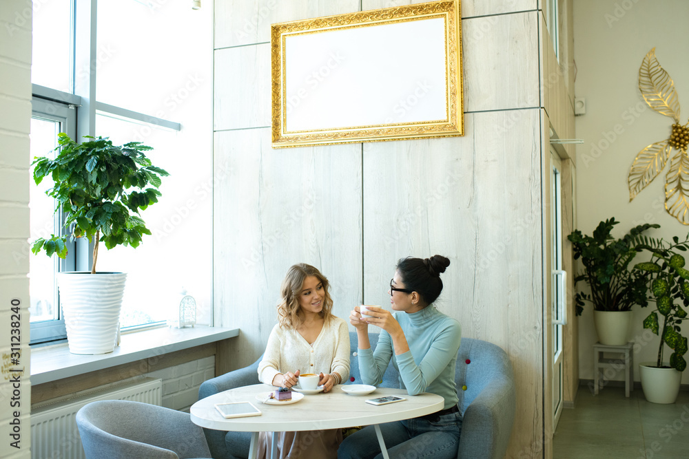 Two friendly young women in casualwear having coffee on soft comfortable sofa Two friendly young women in casualwear having coffee on soft comfortable sofa