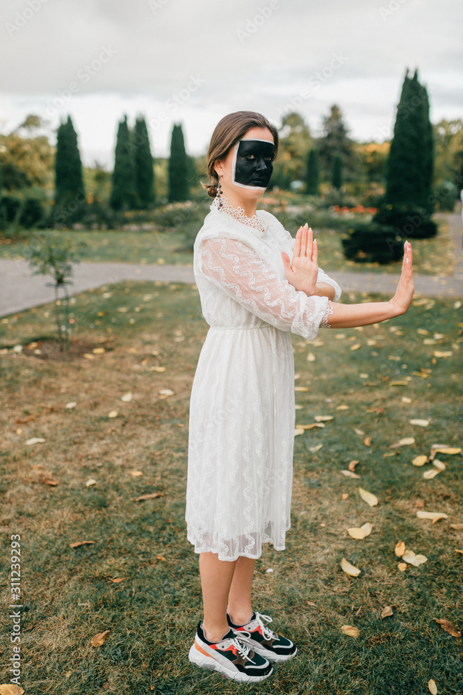 Strange woman in wedding dress with creative face art (black square)showing gestures by her hands in summer park.