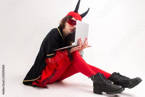 Sexy brunette woman in red tights, black boots and devil horns using laptop over white studio background. photo