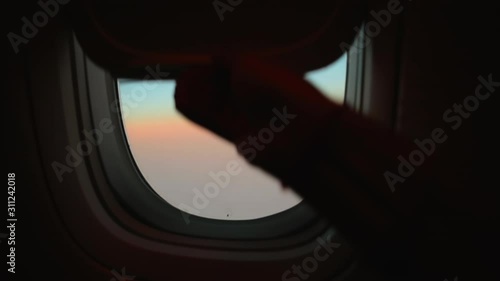 Hand opens airplane window to looks out during air travel. Passenger pov through plane window flight during sunset or sunrise. Close up of porthole of flying airplane at night. Aircraft above clouds.