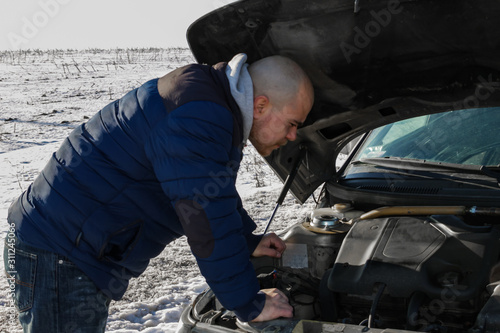 Young man looking under the hood of broken car in cold winter day