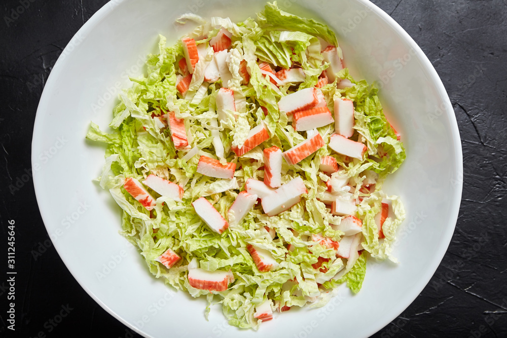 Chopped crab sticks and fresh chinese cabbage leaves in white ceramic bowl on black stone background, top view. Vegetable and imitation crab meat, sea food.  Cooking salad