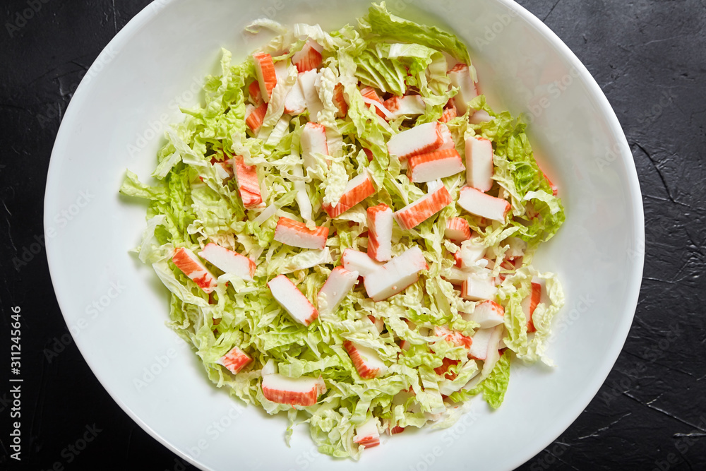 Chopped crab sticks and fresh chinese cabbage leaves in white ceramic bowl on black stone background, top view. Vegetable and imitation crab meat, sea food. Cooking salad Chopped crab sticks and fresh chinese cabbage leaves in white ceramic bowl on black stone background, top view. Vegetable and imitation crab meat, sea food. Cooking salad
