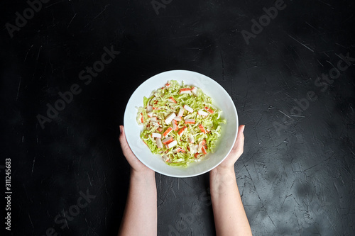 Obraz na plátně Chopped crab sticks and fresh chinese cabbage leaves in white ceramic bowl on black stone background, top view