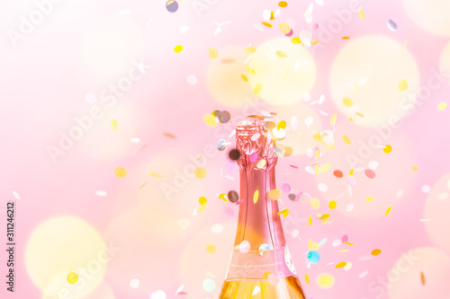 A bottle of sparkling wine with confetti and bokeh in the foreground, on pink background