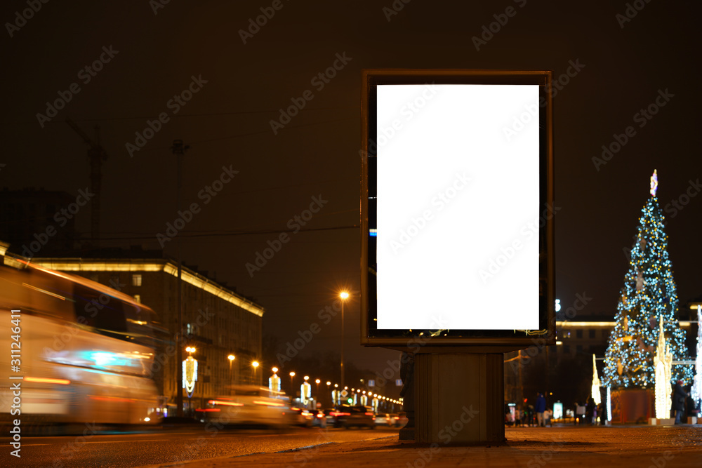 Mockup vertical billboard standing in city city format. Glowing at night billboard with an advertising field. Against the backdrop of a New Year festive tree in the distance blurred.