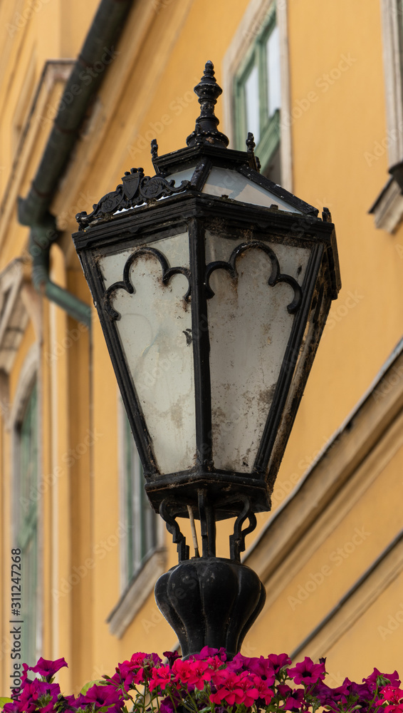 Old iron lamp post with flowers on it. Yellow building in the background