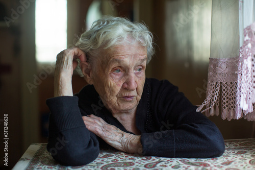 Dramatic portrait of an old gray-haired woman sitting at a table in house..