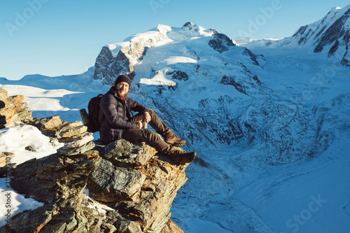 Traveler Man with backpack trekking in mountains, enjoy beautiful Matterhorn view. Explorer man hiking on hills, travel in Swiss Alps, Switzerland. Hiker sitting on rock cliff outdoors on nature.