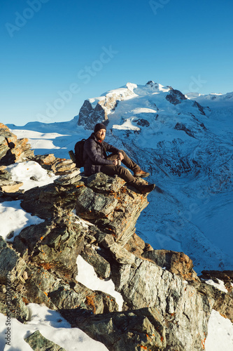 Traveler Man with backpack trekking in mountains, enjoy beautiful Matterhorn view. Explorer man hiking on hills, travel in Swiss Alps, Switzerland. Hiker sitting on rock cliff outdoors on nature.