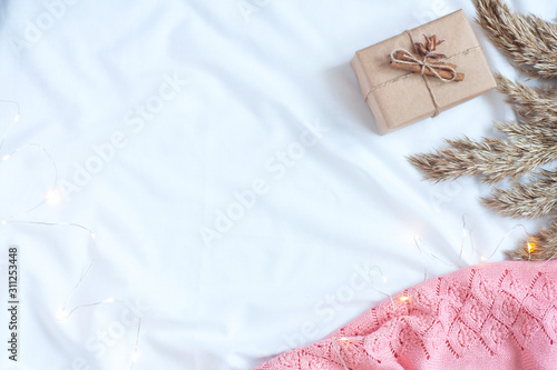 Autumn or winter cozy background. Pink plaid, gift box, dry grass and lights on a white bedding. Home relaxation concept. Flat lay, top view, copy space.