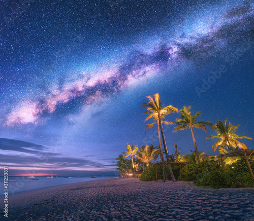 Fototapeta Naklejka Na Ścianę i Meble -  Milky Way over the sandy beach with palm trees and sunbeds and umbrellas at night in summer. Landscape with sea shore, beautiful starry sky, galaxy and green palms. Travel in Zanzibar, Africa. Space