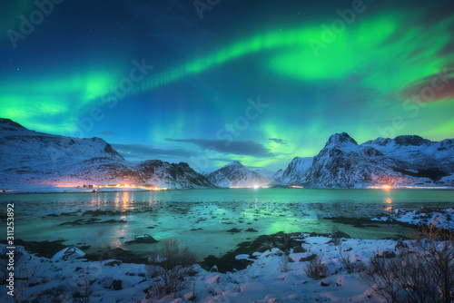 Aurora borealis over the sea coast, snowy mountains and city lights at night. Northern lights in Lofoten islands, Norway. Starry sky with polar lights. Winter landscape with aurora reflected in water © den-belitsky