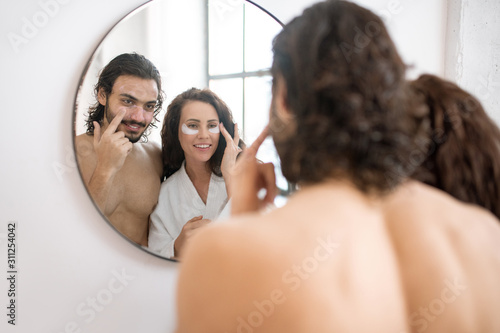 Happy couple taking care of undereye area while standing in front of mirror