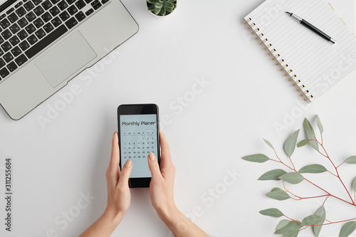 Hands of female employee holding smartphone with monthly planner on screen