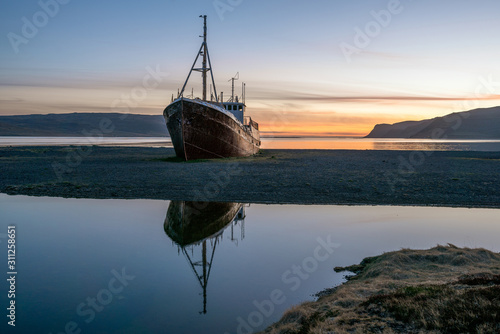 Old ancient, destroyed and abandoned norwegian whaling ship laying ashore in the westfjords of Iceland during sunset. Boat reflects in water. Shipping, whaling and icelandic traveling concept.