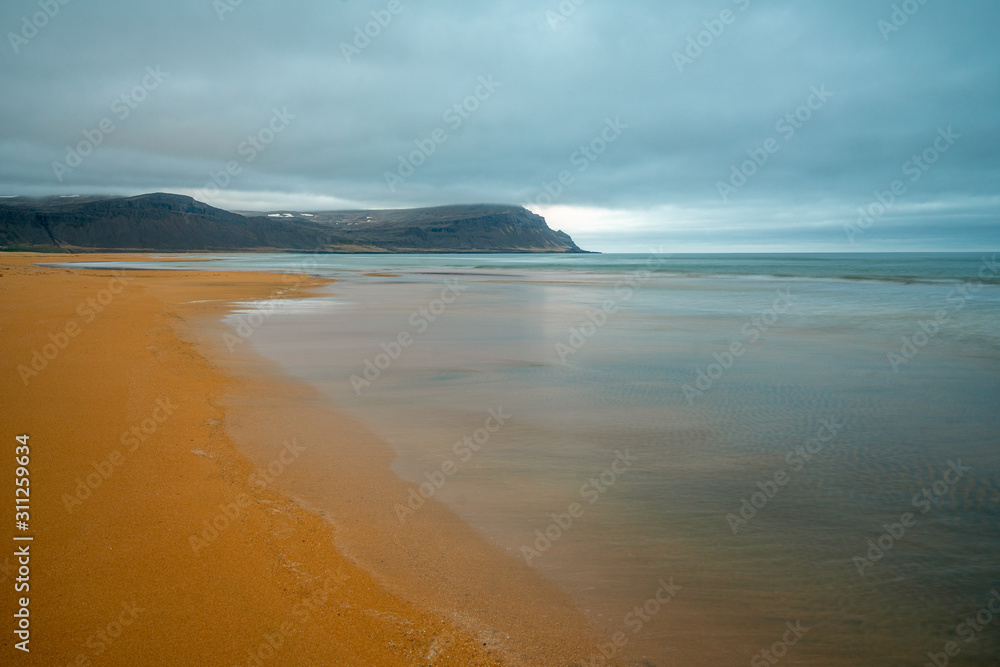 Raudasandur or red sandy beach in the westfjords of Iceland during blue hour. Waves crash into the red sandy dunes at low tide. Texture in the foreground and mountains in the background. Raudasandur or red sandy beach in the westfjords of Iceland during blue hour. Waves crash into the red sandy dunes at low tide. Texture in the foreground and mountains in the background.
