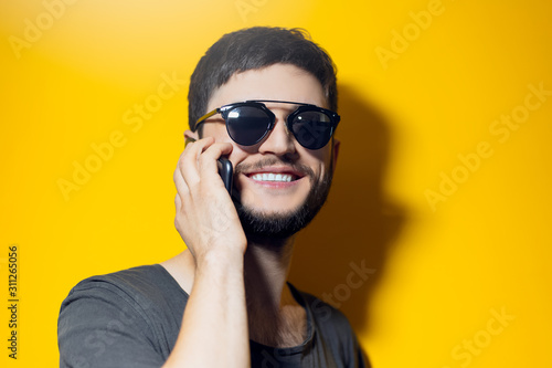 Studio portrait of young smiling man using smartphone, wearing sunglasses on yellow background.