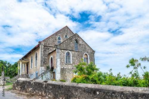 Small modest old stone wall, red zinc roof, stained glass window chapel on rural countryside hill. Rio Bueno Baptist Church in Trelawny parish, Jamaica. Christianity is a dominant Caribbean religion.