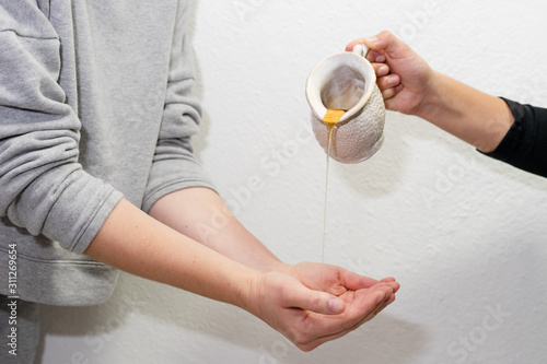 Midsection of two unknown women holding a white ceramic pitcher with raw honey pouring to another woman's female hands in front of the wall