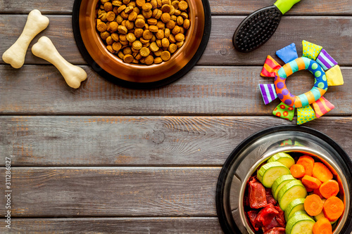 Dog feed - dry food and raw meat - near toy, brush and cheving bones ondark wooden background top view copy space