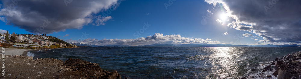 Perspective view of the lake Nahuel Huapi and the cityscape of Bariloche in Argentina in a sunny day, with snowed mountains in the background, seen from stony coast.