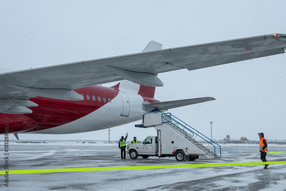 Fototapeta premium Passenger plane at airport in winter in blizzard.A gangway for landing air passengers at the base of a car pulls up to the door of an airplane. A team of airport service people in workwear.