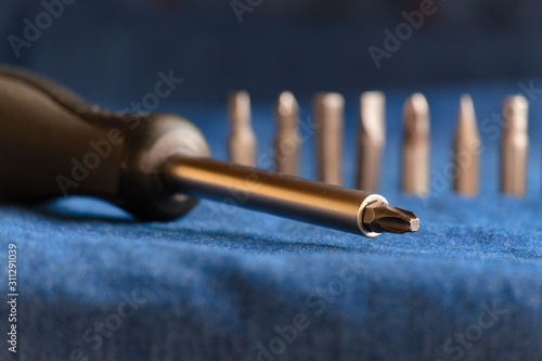 Close-up of stainless steel screwdrivers with black handle and various screwdriver heads placed on a blue denim background.