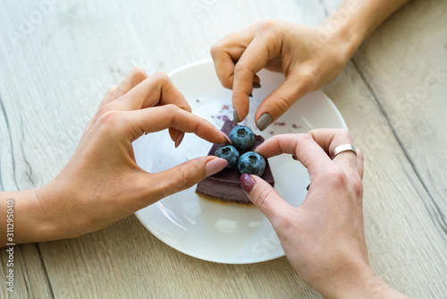 Hands of two girls taking blueberries from top of tasty cheesecake on plate