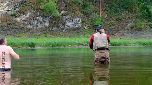 Fly fishing. A photographer with a naked torso waist-deep in water photographs a fisherman. in red clothes. Sports and entertainment on a mountain river.