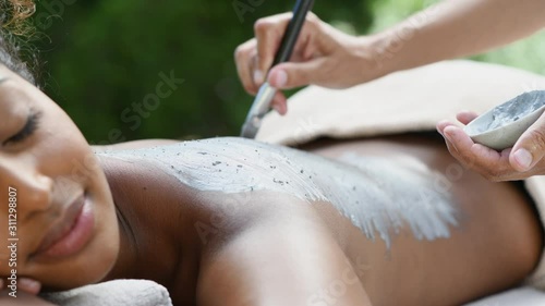 Hands of masseuse applying mud mask on african woman back. Young african american girl relaxing with eyes closed while beautician applying exfoliation clay on shoulder. Woman at resort doing clay mask photo