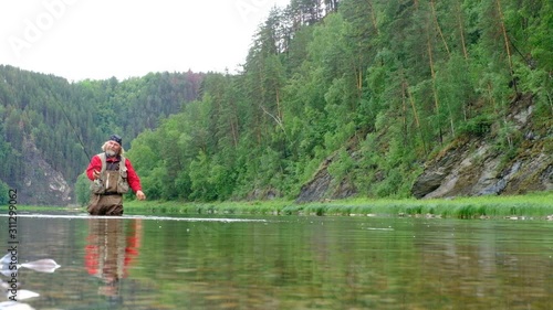 A fisherman in red clothes throws bait into the water with a fishing rod. Fly fishing. Sports and entertainment on a mountain river. Overall plan. Nature, flora and fauna.