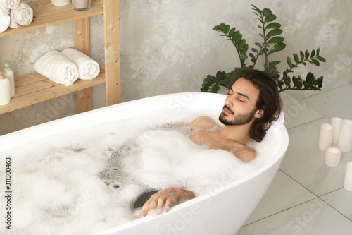 Bild auf Leinwand Young relaxed man with closed eyes lying in bathtub with hot water and foam