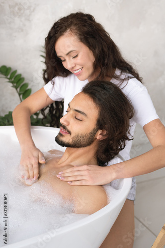 Young smiling woman making massage of chest to her husband relaxing in bath