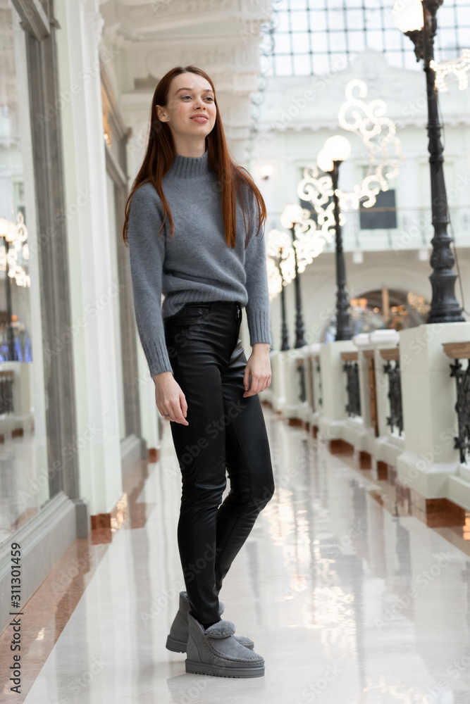 European tall girl long red hair in leather pants, gray uggs and a sweater posing in a gallery near boutiques Stock Photo | Adobe Stock