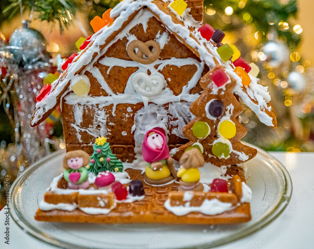 Selective focus on homemade and home decorated ginger bread house with soft focus sugar candy characters out front on a white table in front of a decorated Christmas tree