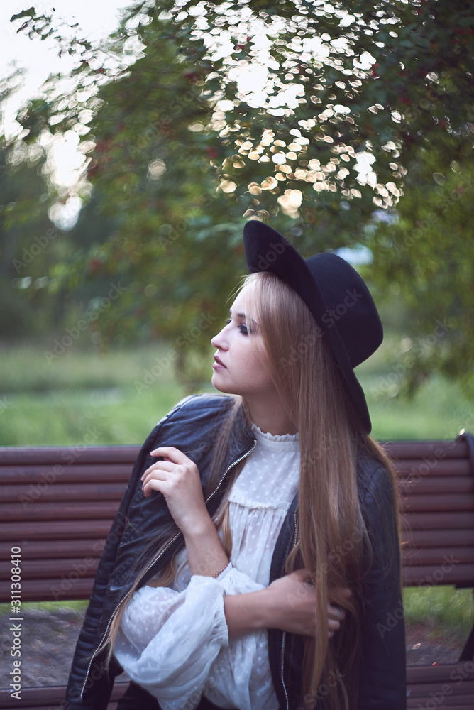 Naklejka premium A European white woman in a black hat and white shirt sits on a wooden bench. Summer portrait