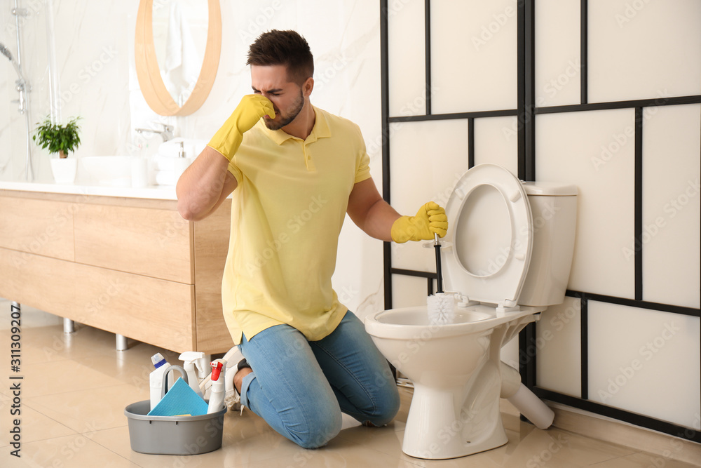 Young man feeling disgust while cleaning toilet bowl in bathroom - obrazy, fototapety, plakaty 