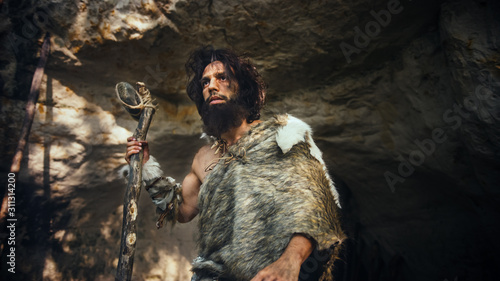 Primeval Caveman Wearing Animal Skin Holds Stone Tipped Hammer Comes out of the Cave and Looks Around Prehistoric Forest, Ready to Hunt Animal Prey. Neanderthal Going Hunting into the Jungle.
